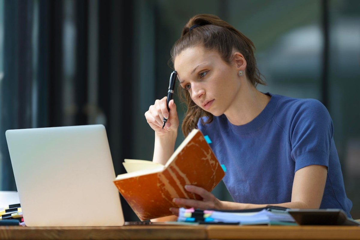 A woman in a blue shirt takes notes in a notebook while seated at a wooden table with a laptop and colorful stationery.