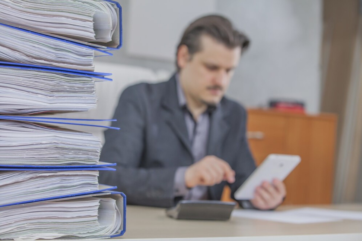 A stack of blue folder paper documents stands in focus while a blurred figure works on a tablet at a desk in an office setting.