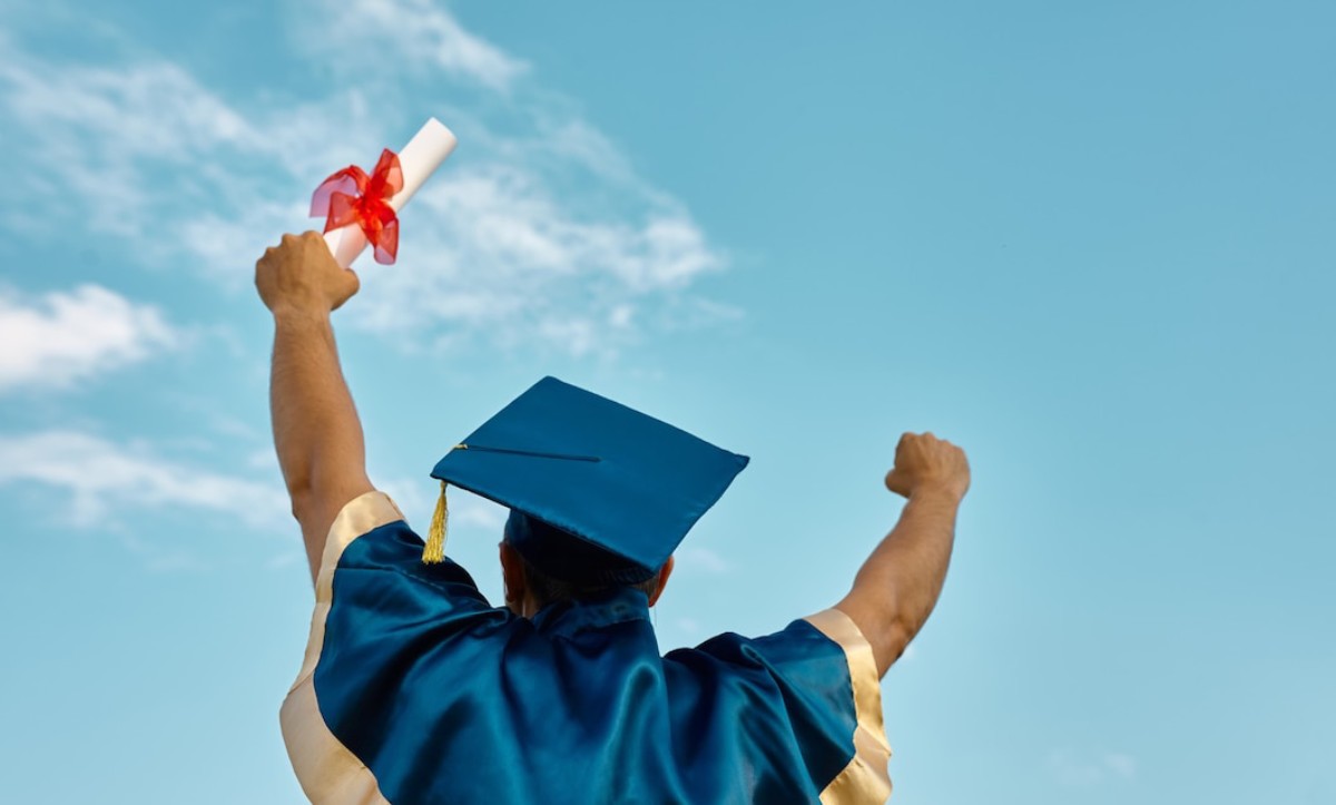 A graduate in a blue cap and gown triumphantly raises a diploma tied with a red ribbon against a bright blue sky.