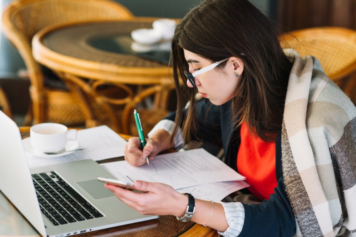 A person sitting at a table, working on a laptop, taking notes, with a cup of coffee and papers scattered around.