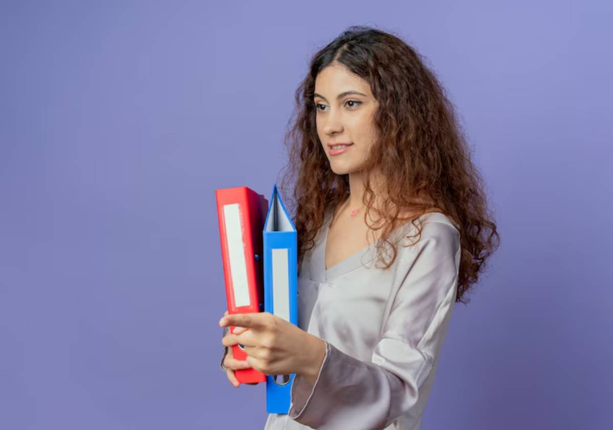 A person with curly hair holds a red and a blue binder, set against a soft purple background.