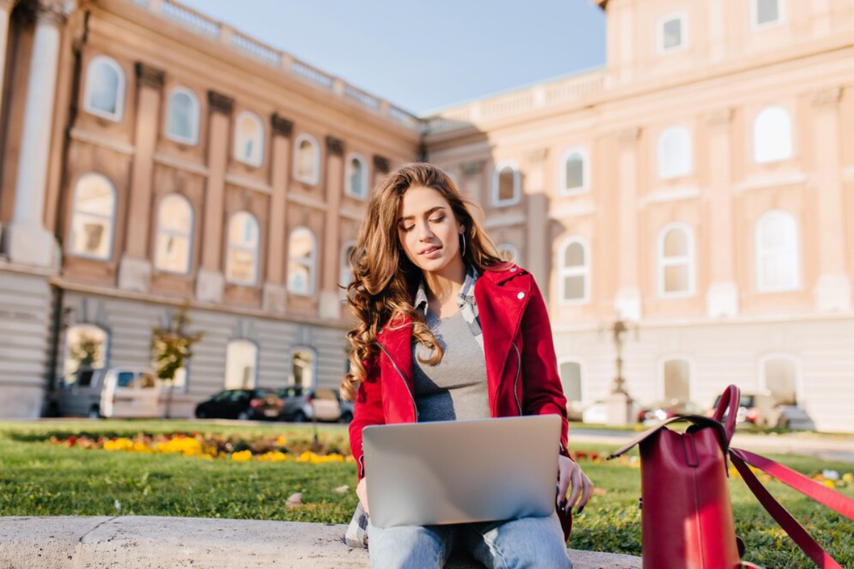 A woman with long, wavy hair sits on a grassy lawn, working on a laptop in front of a historic building. A red handbag lies beside her.