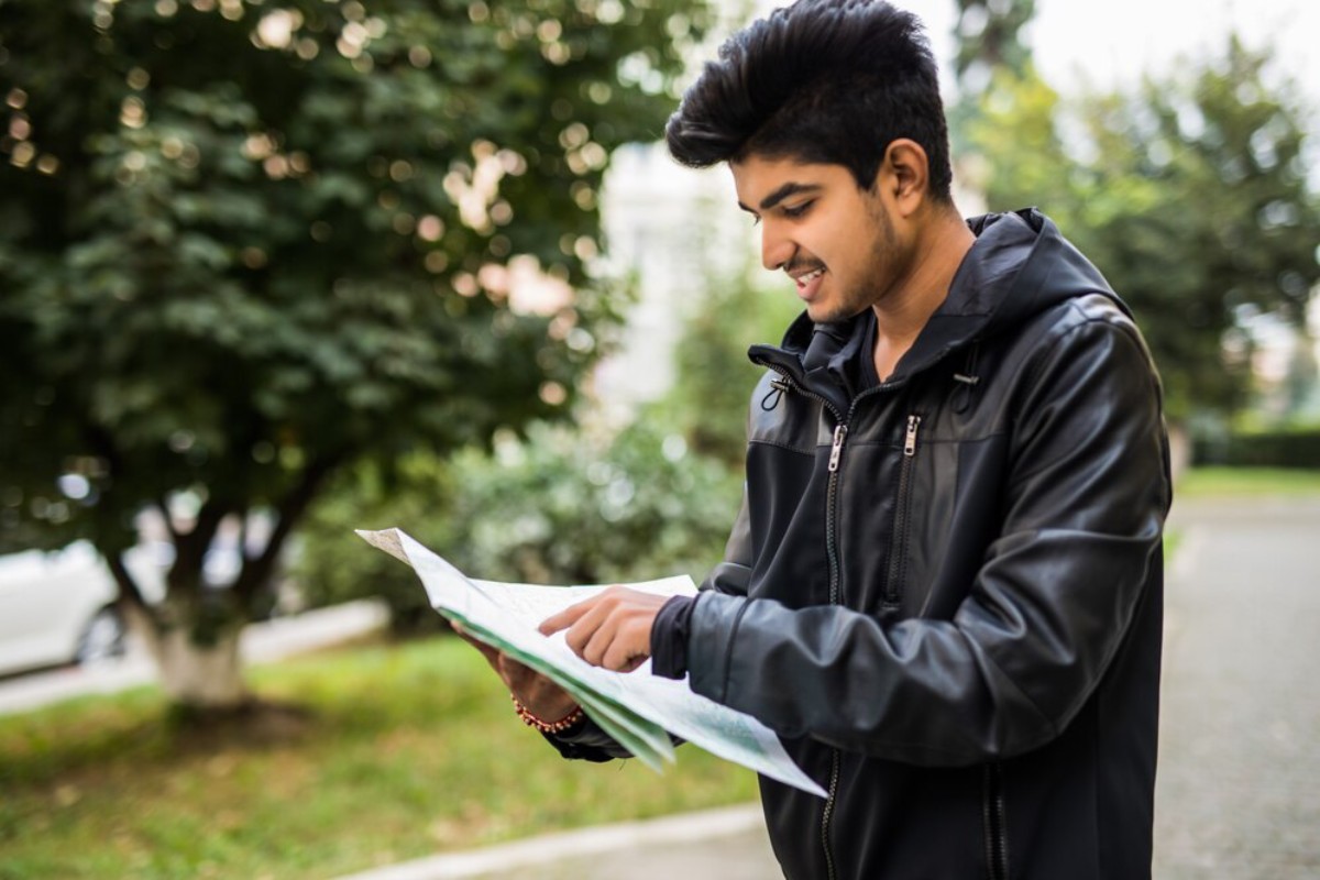 A person in a black jacket is intently studying a map while standing in a green park with trees in the background.