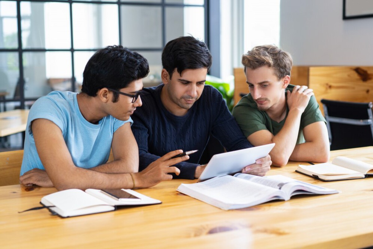Three people are gathered around a tablet, engaged in discussion, with notebooks and textbooks on a wooden table in a bright room.