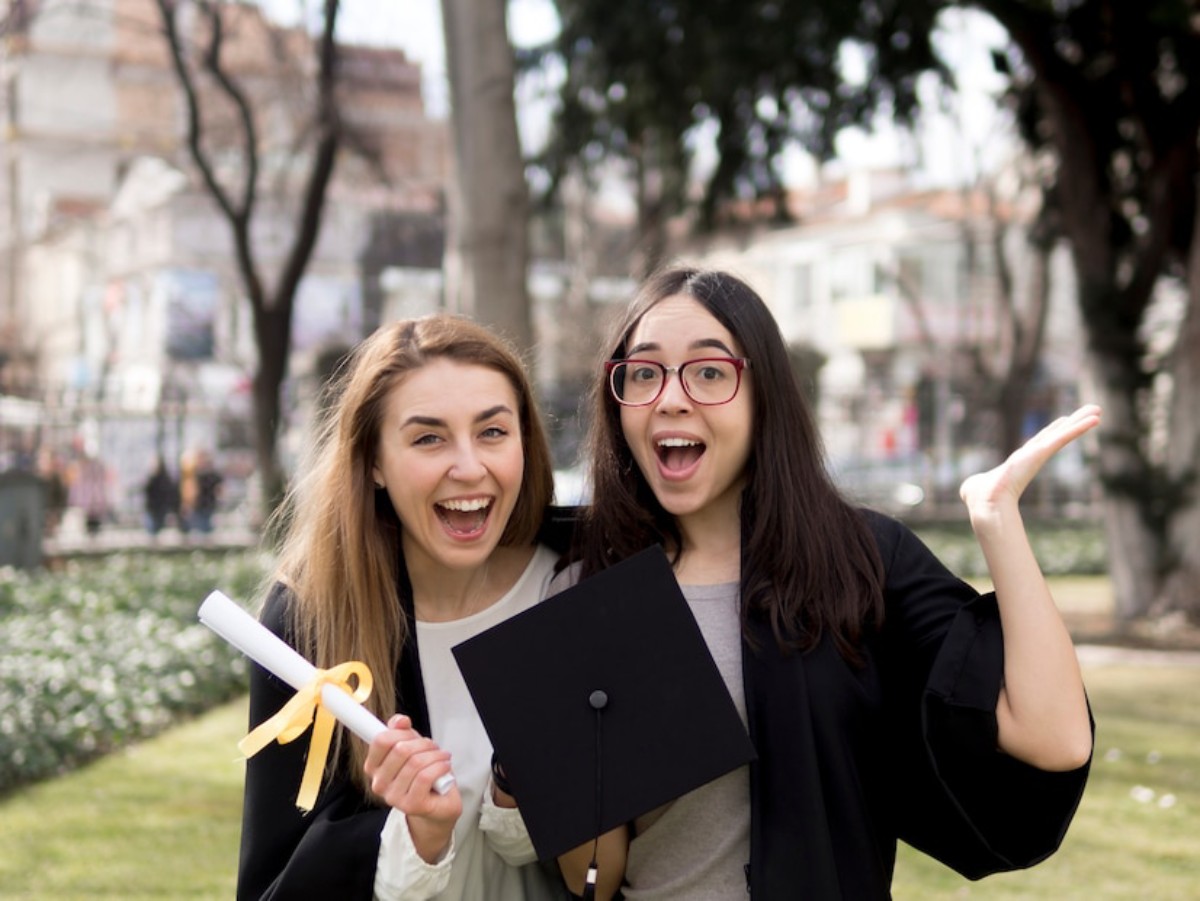 Two graduates in caps and gowns pose joyfully outside with a diploma, celebrating their achievement in a park setting.