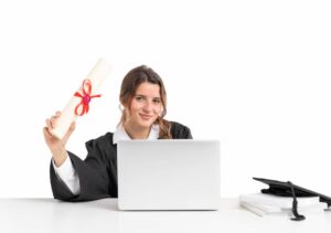 A person in a graduation gown holds a diploma while sitting at a desk with a laptop and graduation cap.