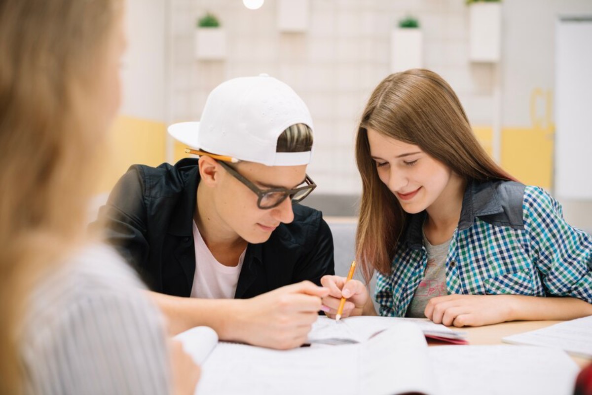 Two students lean over a table, collaborating on a project with books and stationery scattered around them in a bright study space.
