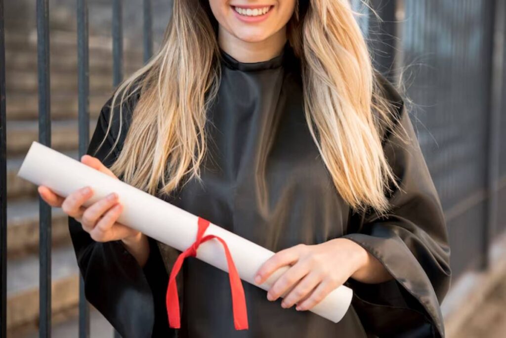 Smiling graduate in a black gown holds a rolled diploma tied with a red ribbon against a blurred outdoor background.