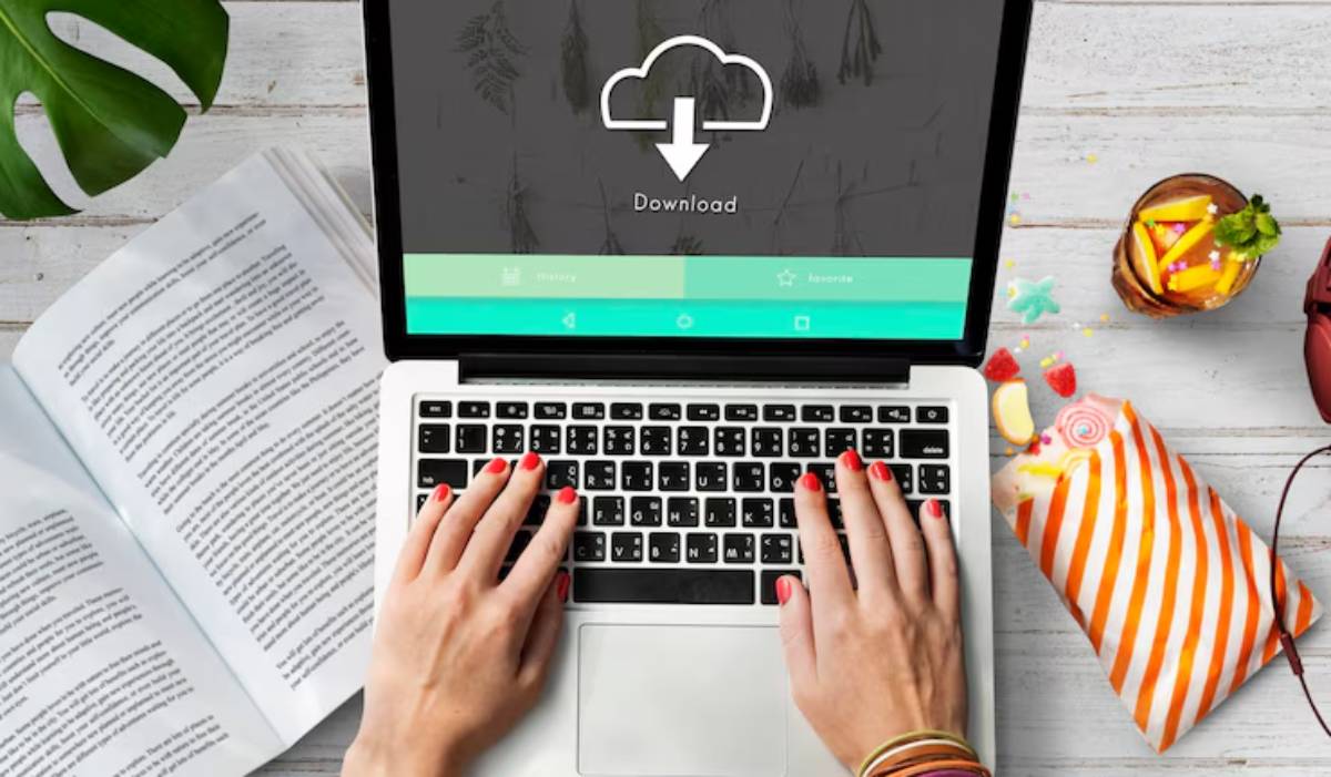 A person with red nails types on a laptop displaying a download icon, surrounded by snacks, a book, and a tropical drink.