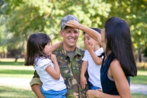 A soldier in camouflage stands in a park, surrounded by two children and an adult woman, all engaged in a joyful family moment.