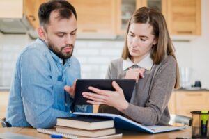 A man and woman sit at a table in a kitchen, discussing content on a tablet, surrounded by books and stationery.