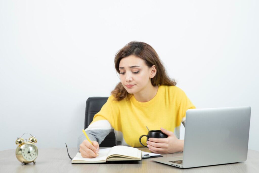 A person in a yellow sweater sits at a desk, writing in a notebook while holding a coffee mug and facing a laptop, with a clock nearby.