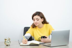 A person in a yellow sweater sits at a desk, writing in a notebook while holding a coffee mug and facing a laptop, with a clock nearby.