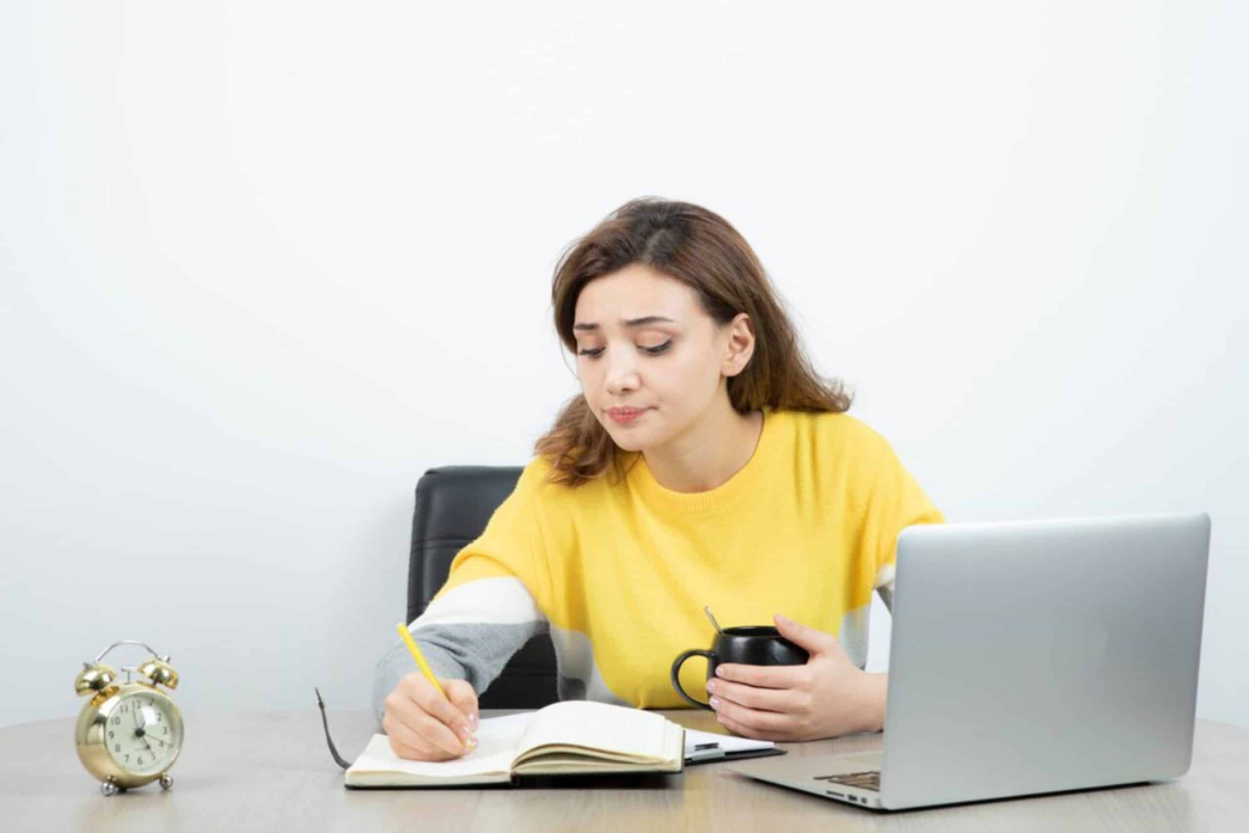 A person in a yellow sweater sits at a desk, writing in a notebook while holding a coffee mug and facing a laptop, with a clock nearby.
