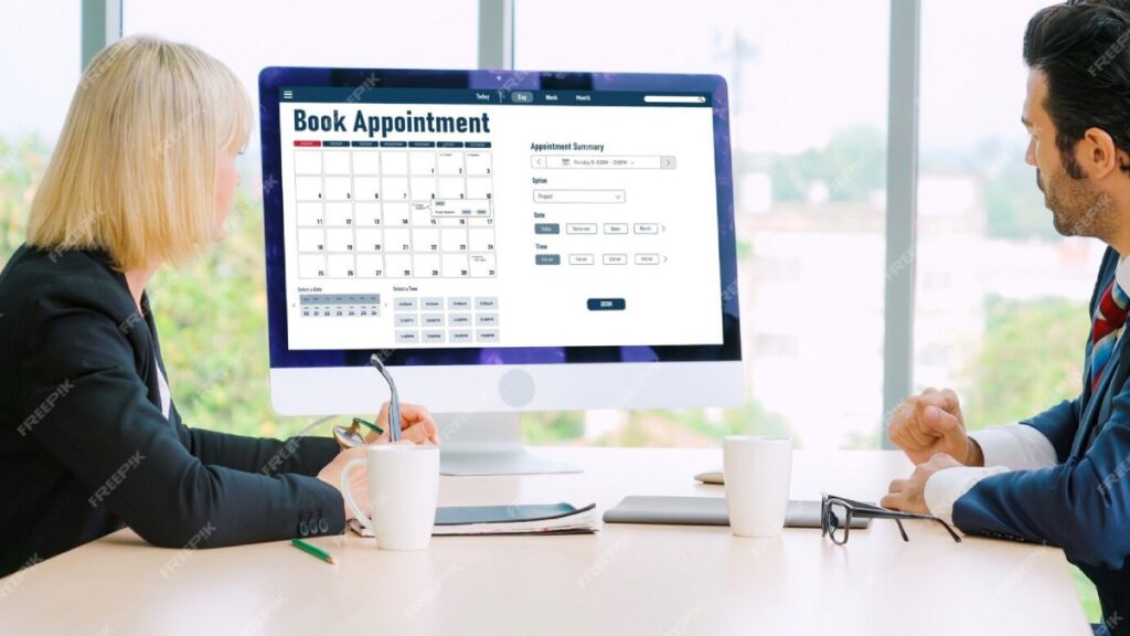 A business meeting taking place with a large computer screen displaying a calendar and appointment booking interface.
