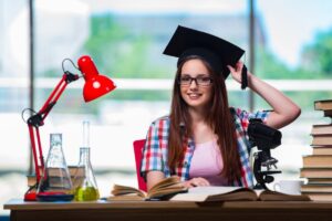 A student in a checkered shirt sits at a desk with books, a microscope, and laboratory glassware, under a red desk lamp.