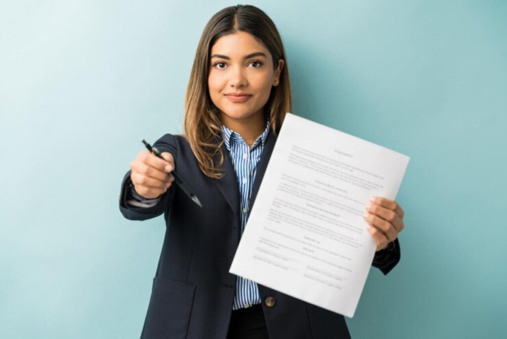 A person in a business suit holds a contract in one hand and a pen in the other against a light blue background.