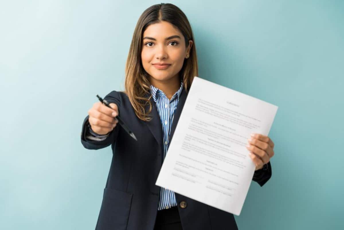 A person in a business suit holds a contract in one hand and a pen in the other against a light blue background.