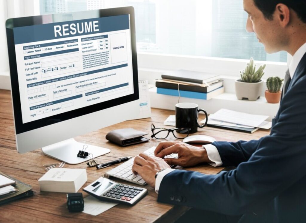A person in a suit types on a computer displaying a resume while sitting at a desk with stationery, a calculator, and coffee.