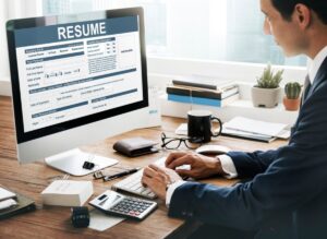 A person in a suit types on a computer displaying a resume while sitting at a desk with stationery, a calculator, and coffee.