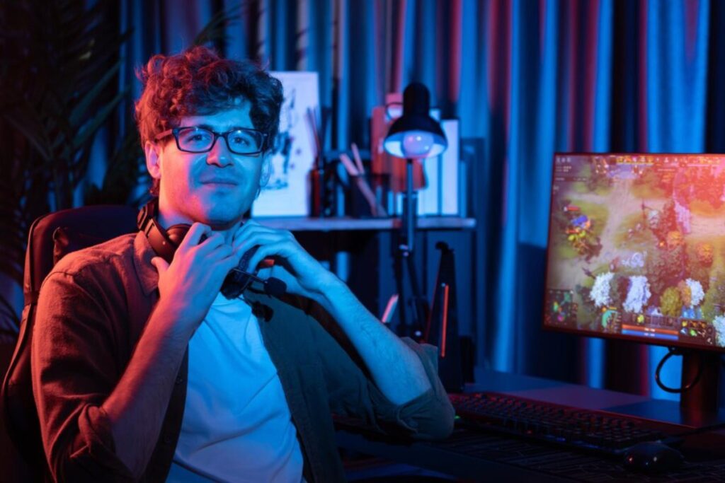 A person sits at a gaming desk, adjusting their headset, with a colorful game displayed on the monitor in a dimly lit room.