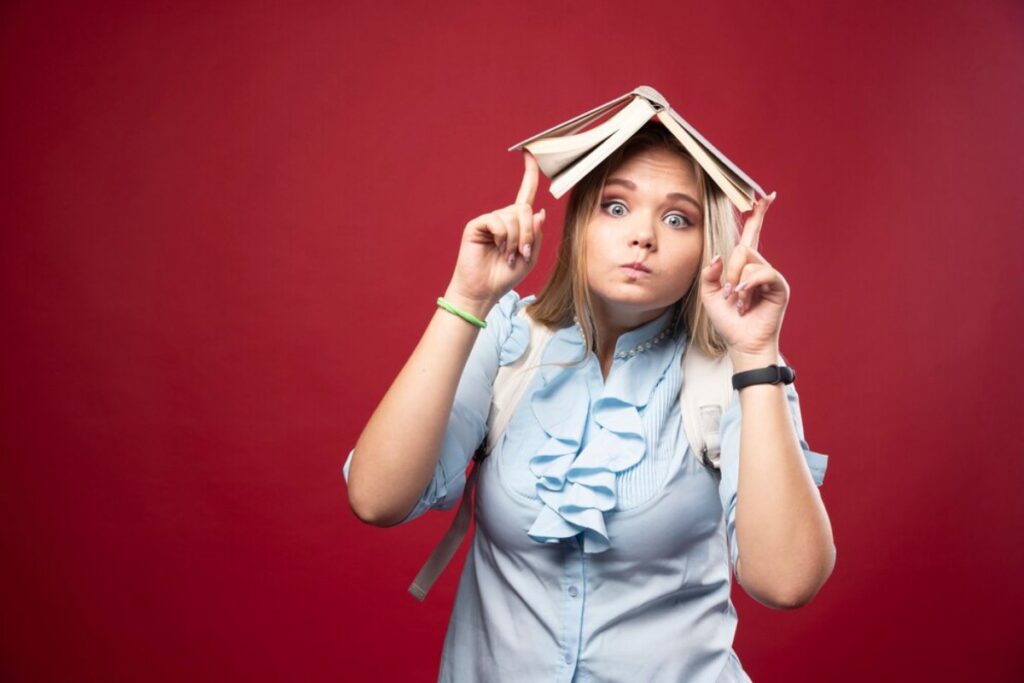 A person wearing a blue shirt and backpack holds a book above their head, with a red backdrop, striking a playful pose.