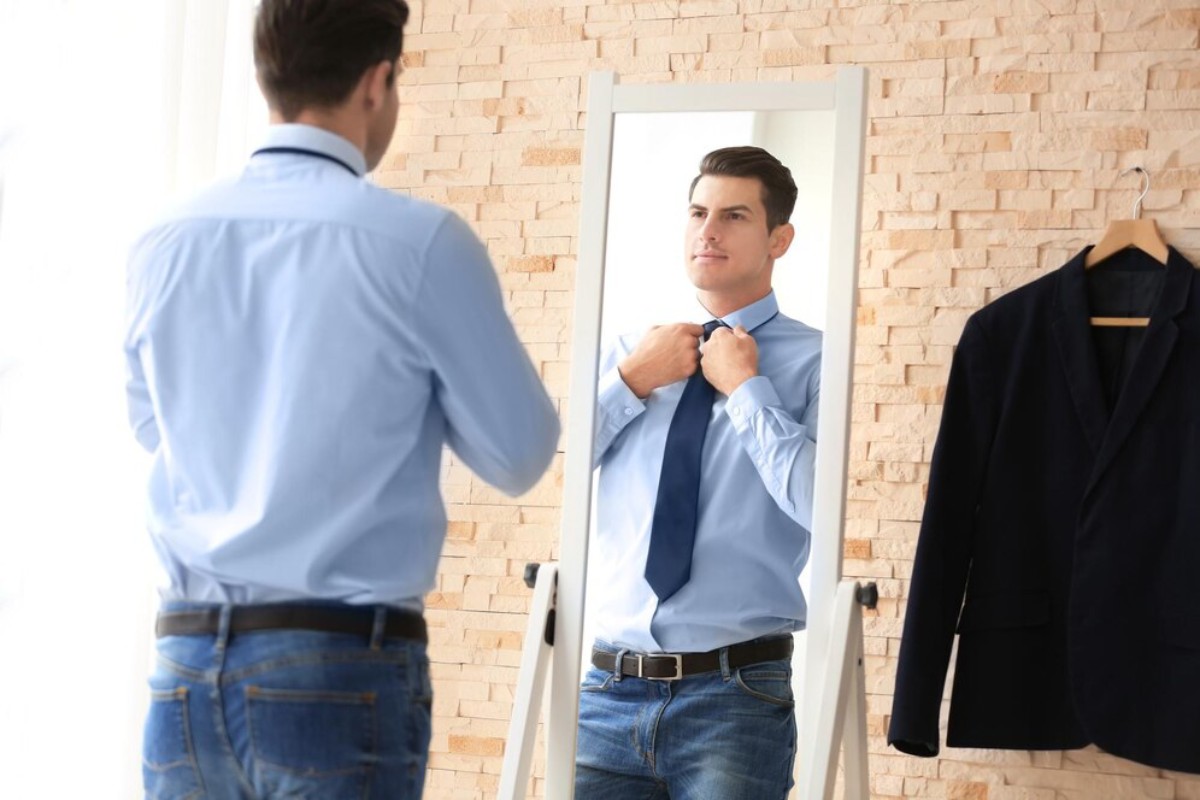 A man in a light blue shirt adjusts his tie in front of a mirror, with a dark jacket hanging nearby. The room has a bright, airy feel.