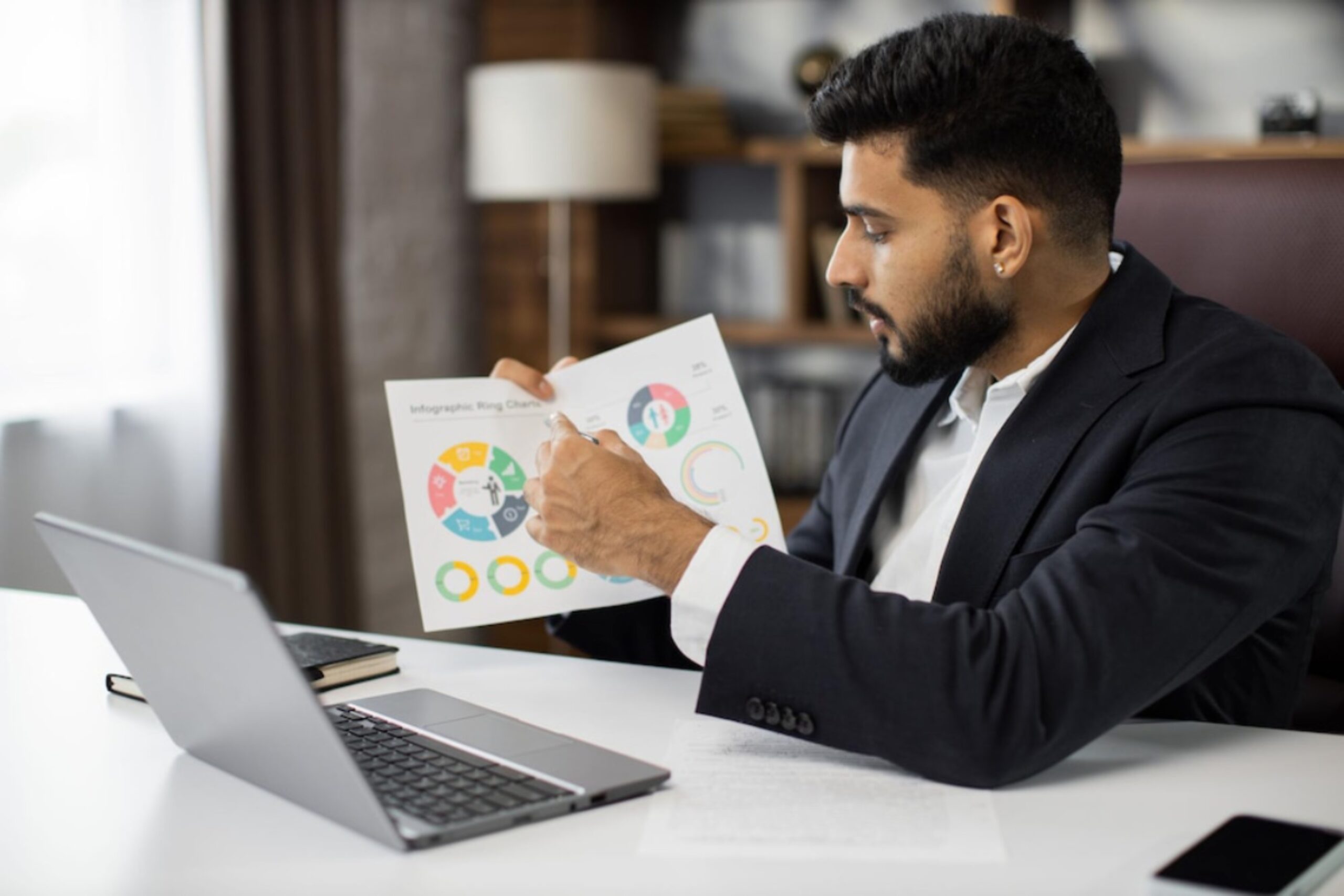 A person in a suit gestures toward an infographic chart while seated at a desk with a laptop and notebook. Natural light floods the room.