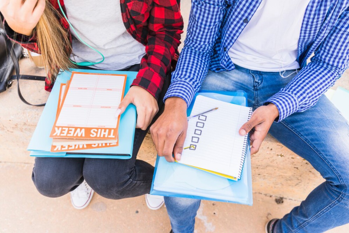 Two students seated outdoors, sharing study materials; one holds a planner labeled 