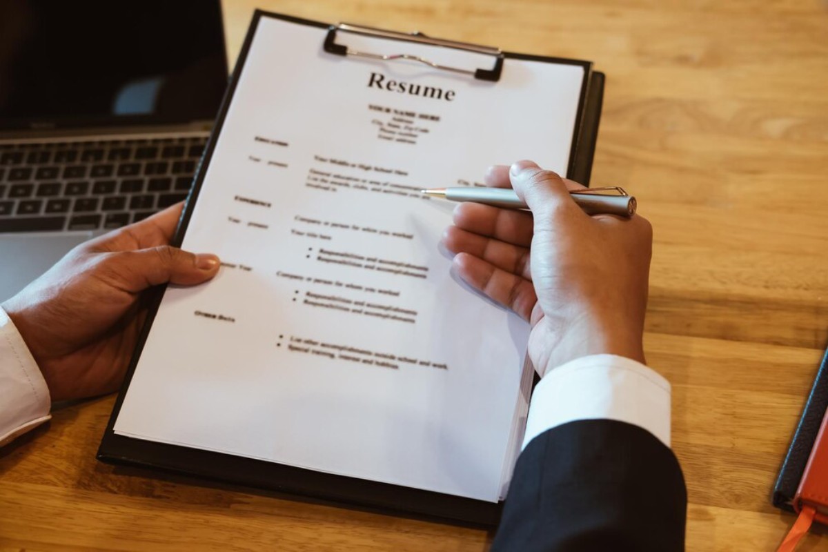 A person in a suit reviews a resume with a pen, seated at a desk with a laptop, focusing on professional details.