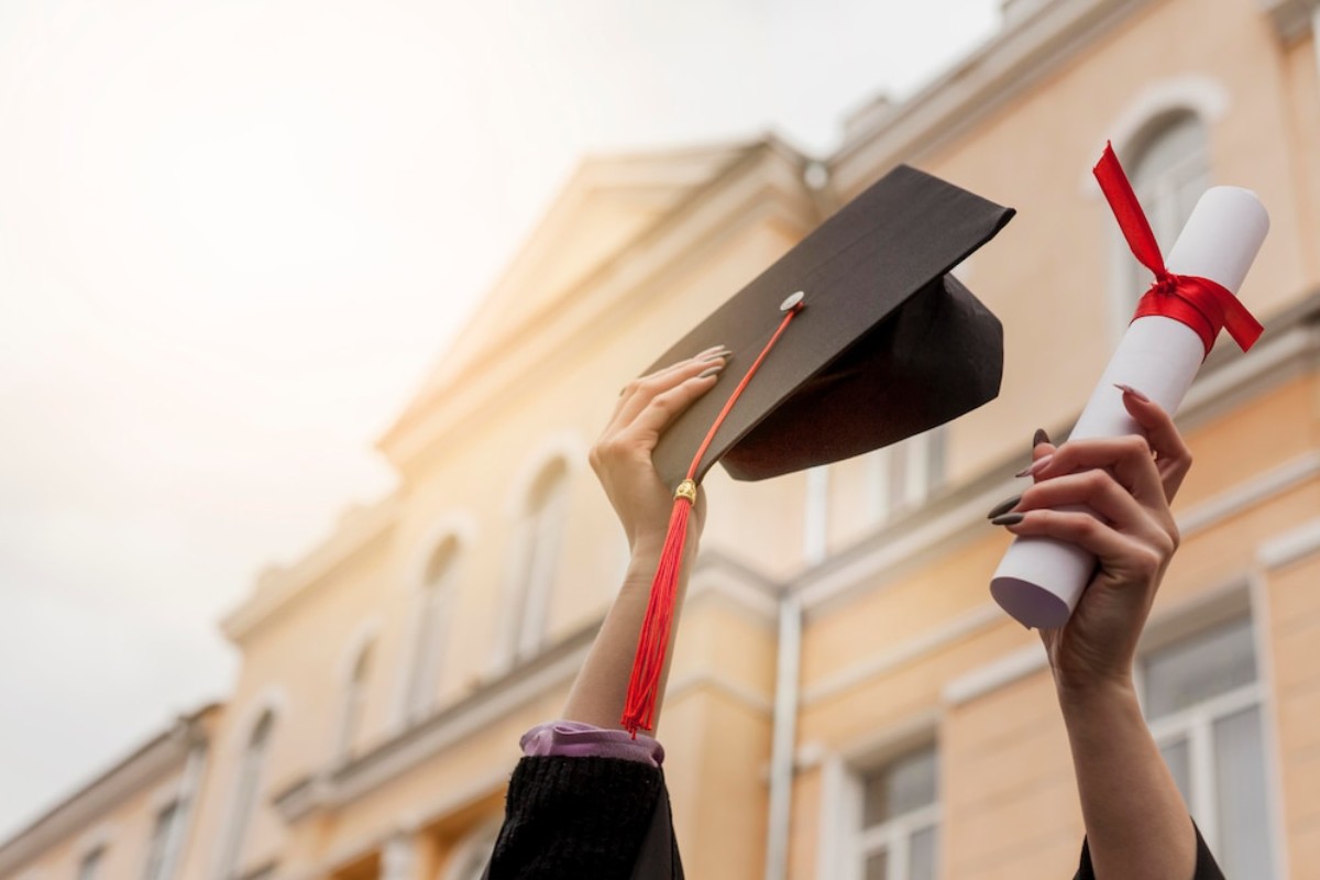 A graduate joyfully raises a mortarboard and diploma in front of a university building, celebrating their achievement.