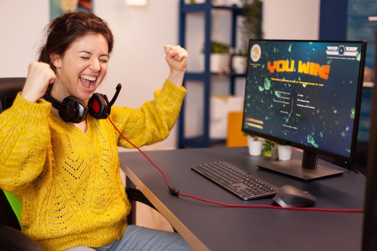 A person in a bright yellow sweater celebrates victory while gaming, sitting at a desk with a monitor, keyboard, and headphones.