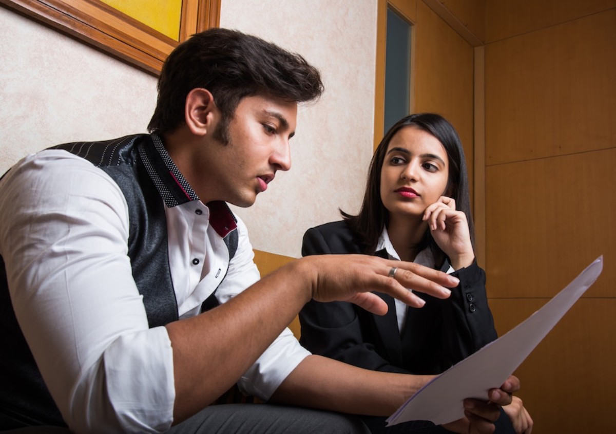 A man in formal attire gestures while discussing a document with a woman in a suit, sitting together in a professional setting.