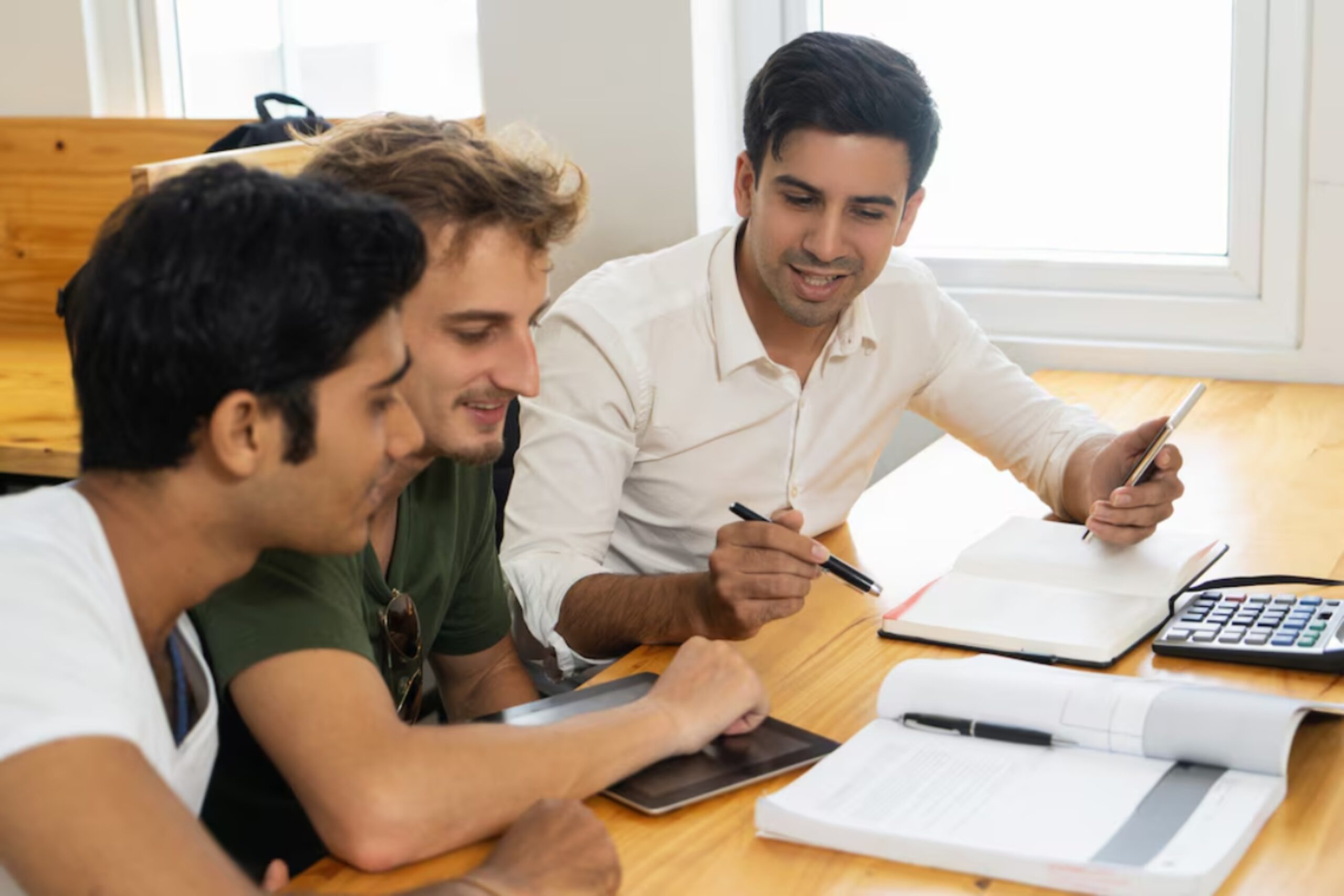 Three individuals engaged in discussion at a wooden table, with notebooks, a calculator, and digital devices in front of them.