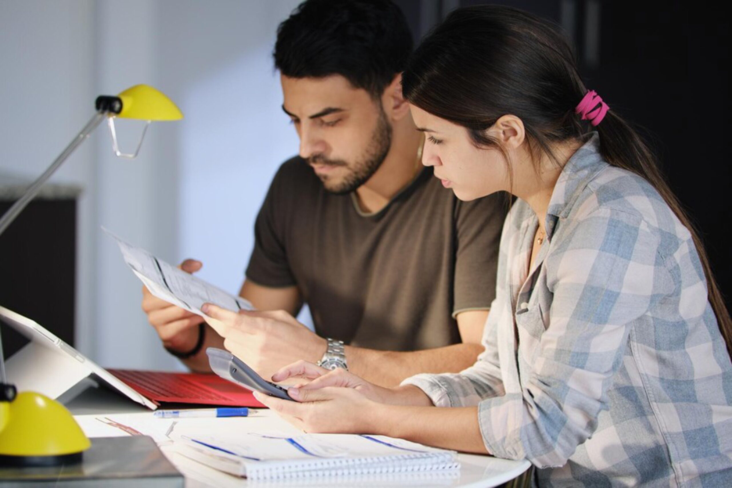Two people collaborate at a table, reviewing documents and using a smartphone under a yellow lamp in a cozy study area.