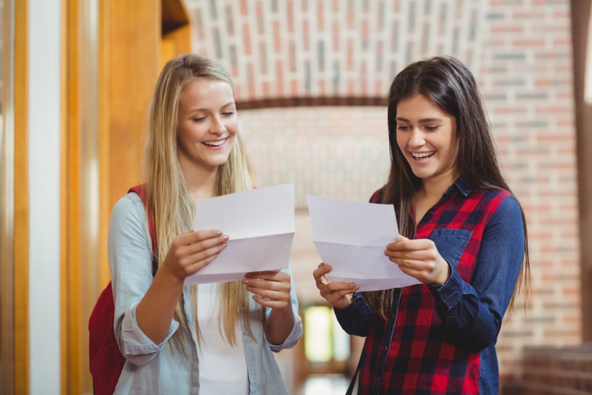 Two young women stand in a hallway, reading letters in their hands, with a rustic brick wall and wooden accents in the background.