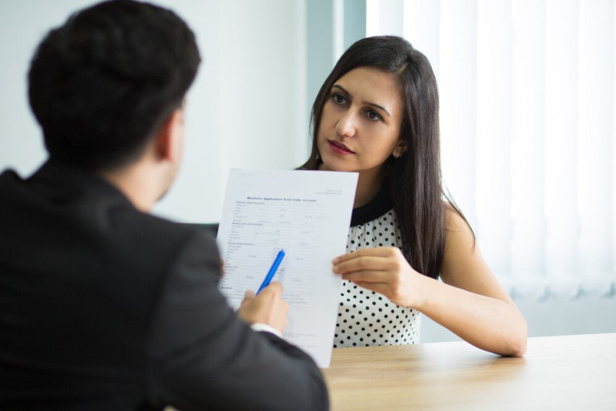 A person in formal attire is seated at a table, reviewing a business application form with a pen in hand. Bright, windowed room.