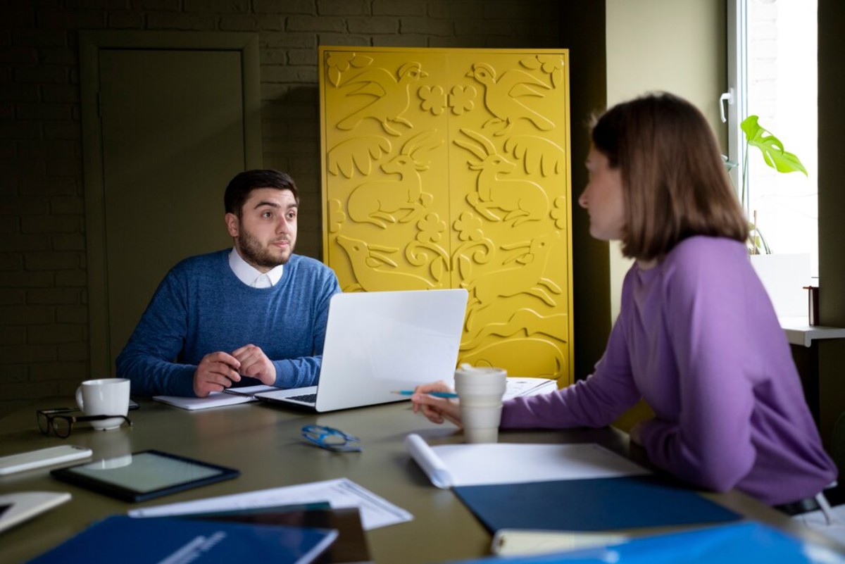 A man in a blue sweater sits at a table with a laptop, while a woman in a purple sweater gestures with a cup in a bright, modern workspace.