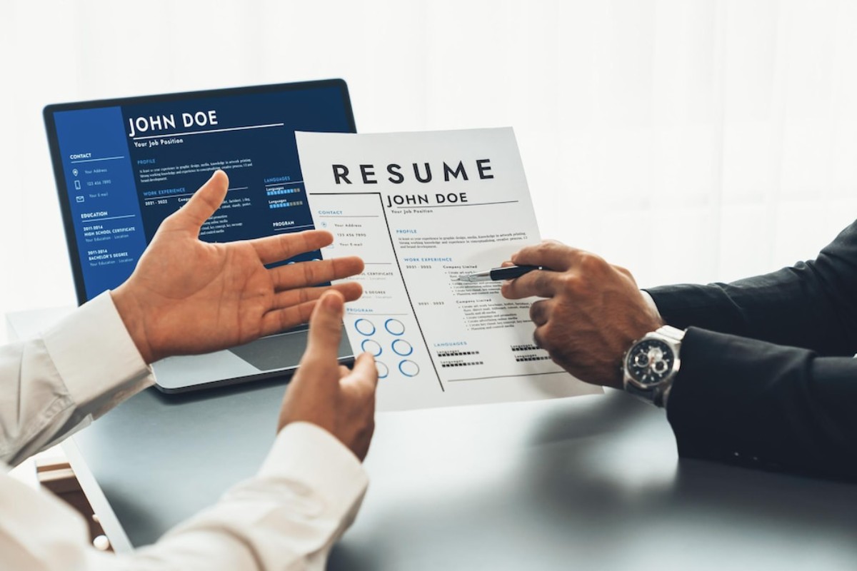 A job interview scene with a candidate presenting their resume to an interviewer, with a laptop displaying the candidate's online profile.