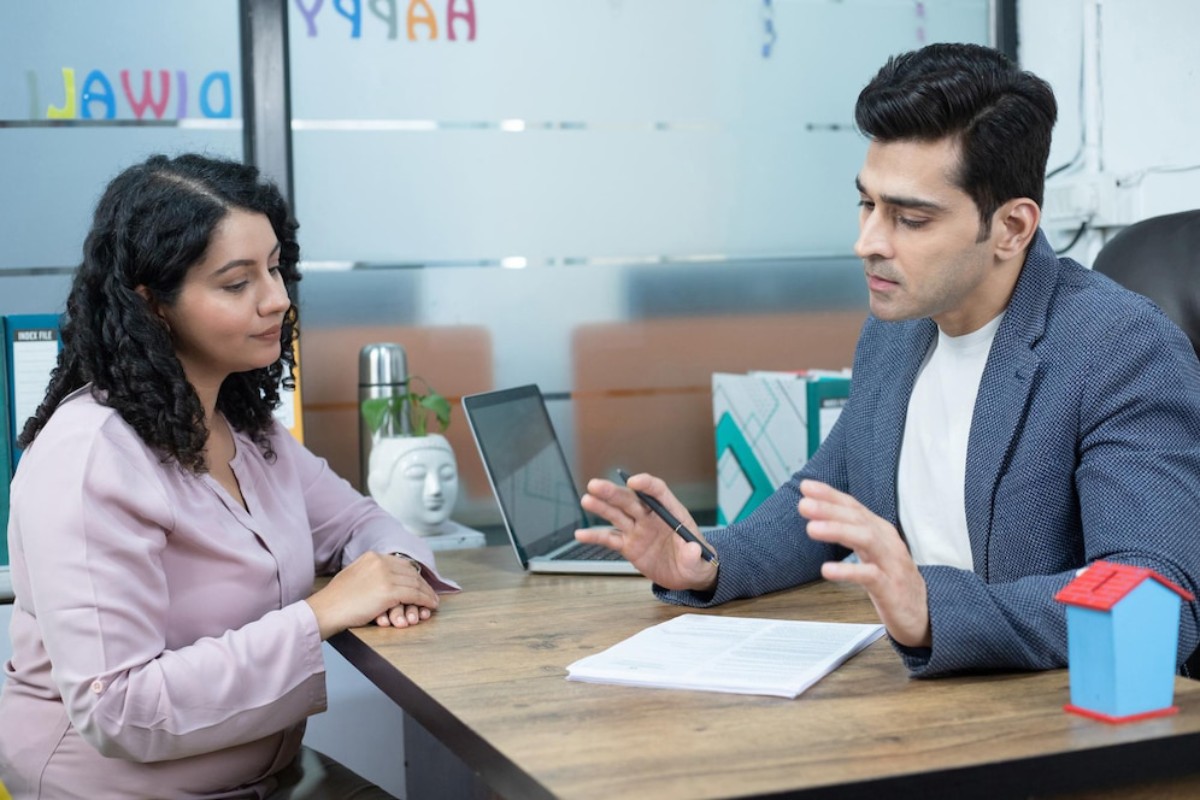 A man in a blazer gestures while discussing with a woman in a pink blouse at an office table with a laptop and documents.