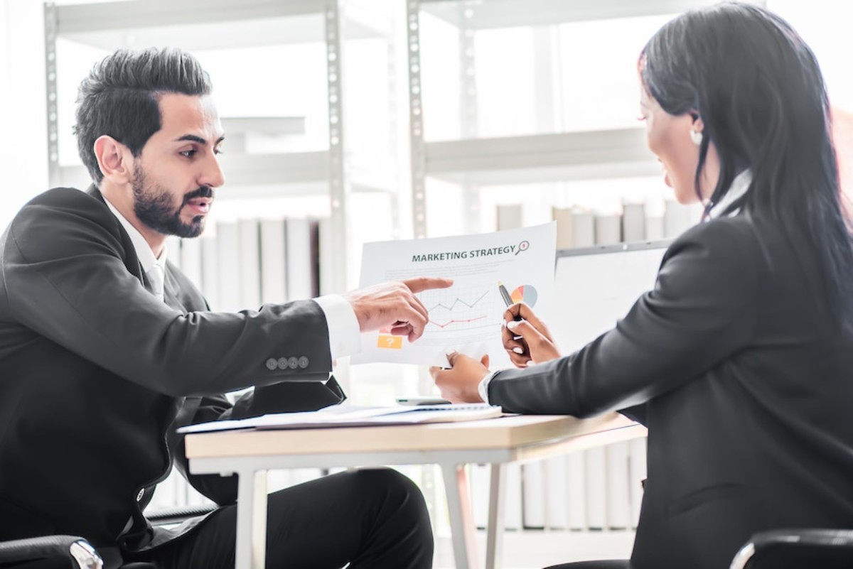 Two professionals in business attire discuss a marketing strategy, analyzing charts and documents at a modern office desk.