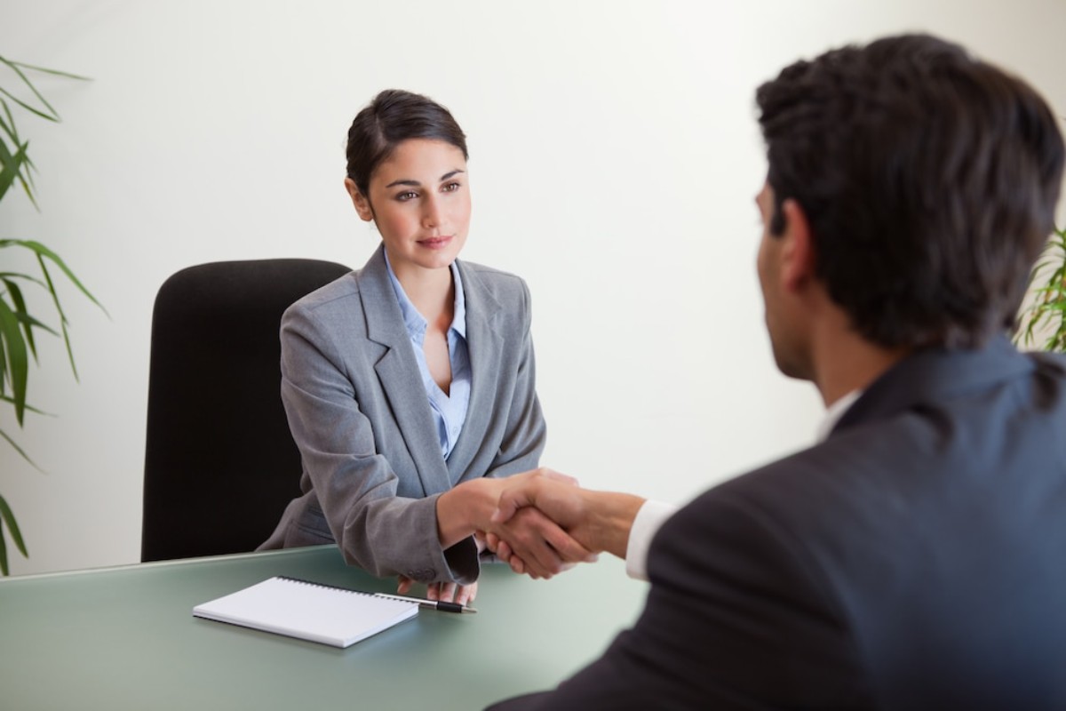 A professional business meeting shows two individuals shaking hands across a desk with a notepad and pen visible. Green plants decorate the background.