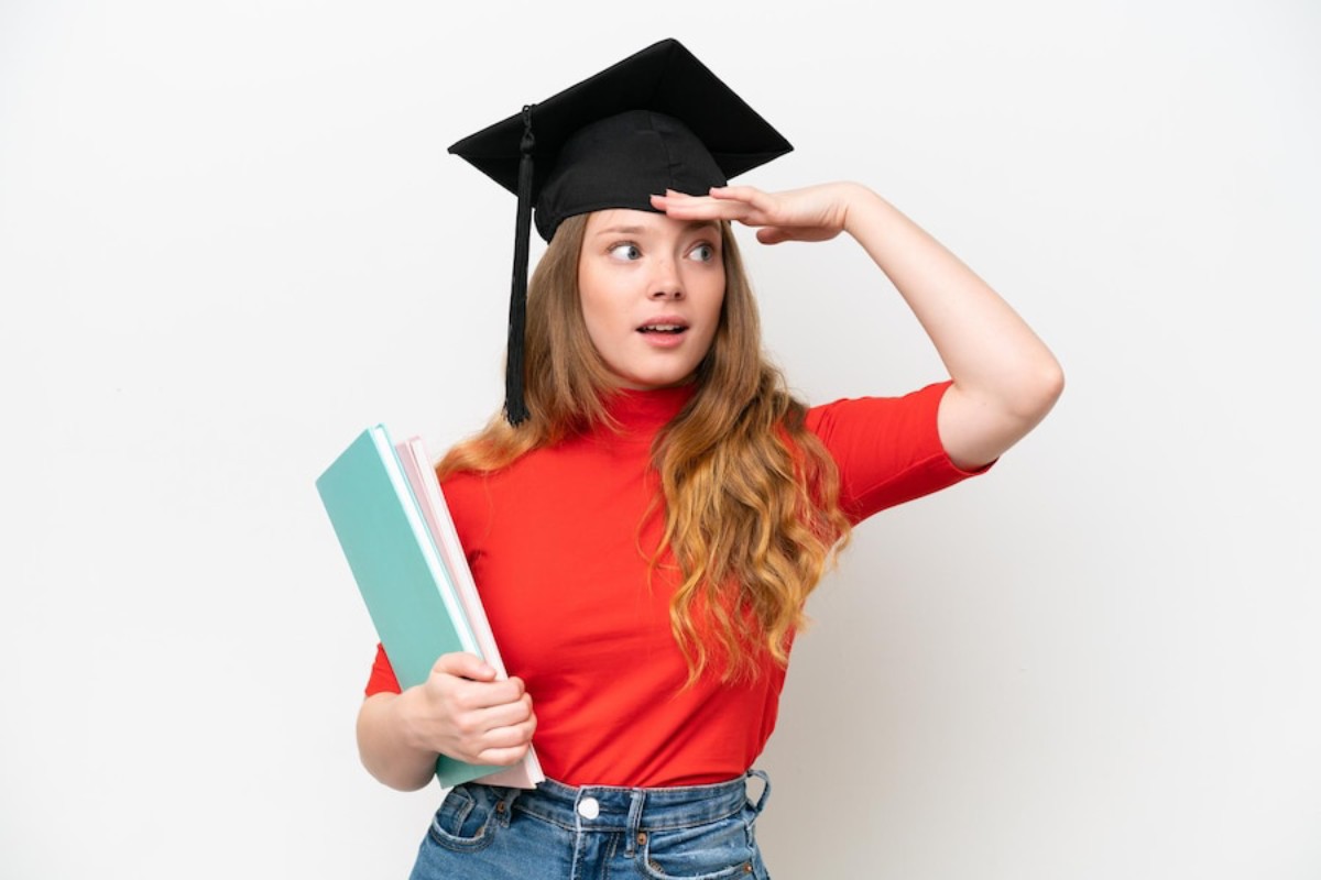 A student in a red shirt and denim shorts strikes a confident pose, wearing a graduation cap and holding books in one hand.