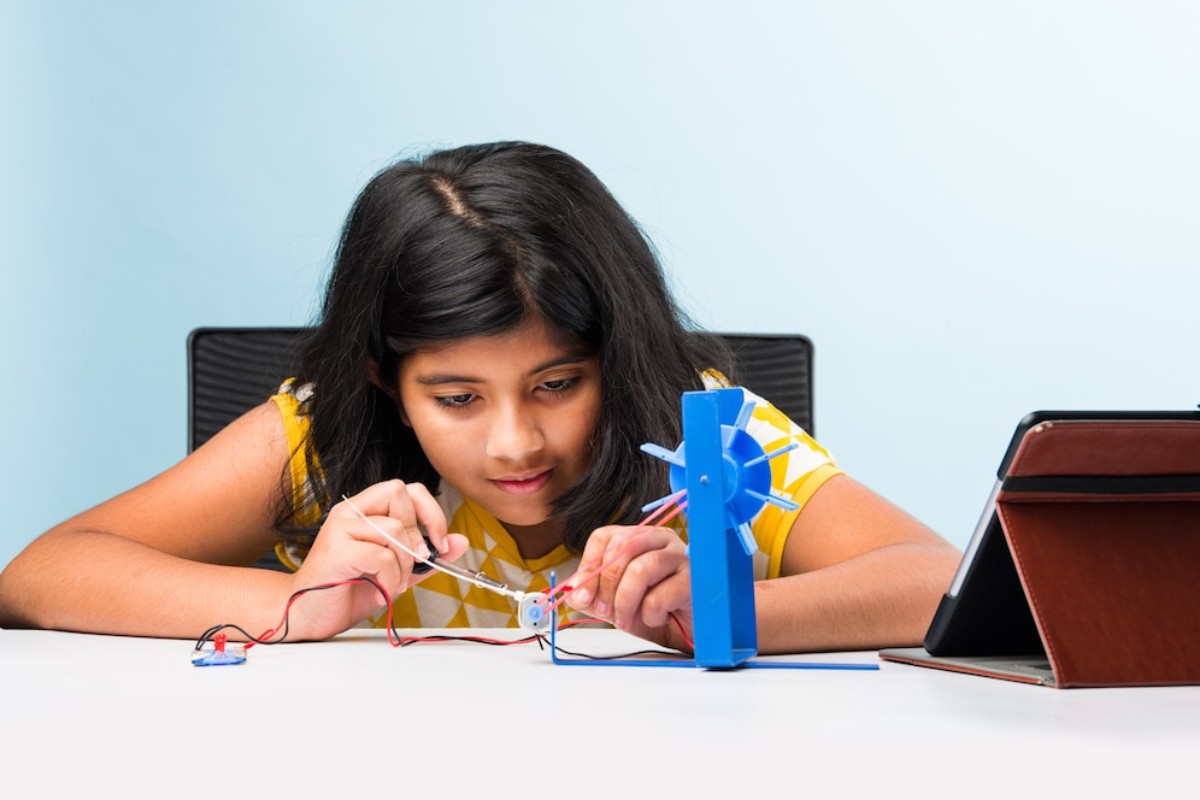A child assembles a small blue fan with wires on a table, focused on their task, with a tablet propped beside them.