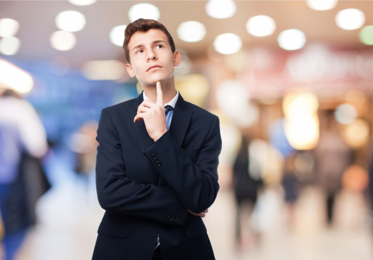 A man in a dark suit poses with a finger to his lips, against a blurry, bustling background of people and soft lights.