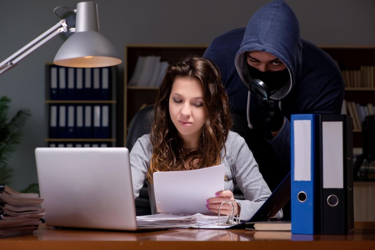 A person in a grey hoodie looms over a seated individual reviewing papers at a desk cluttered with folders and a laptop, creating a tense atmosphere.