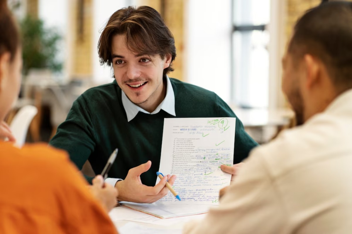 A group discussion at a table, with one person reviewing a marked paper covered in notes and checkmarks, engaged in conversation.