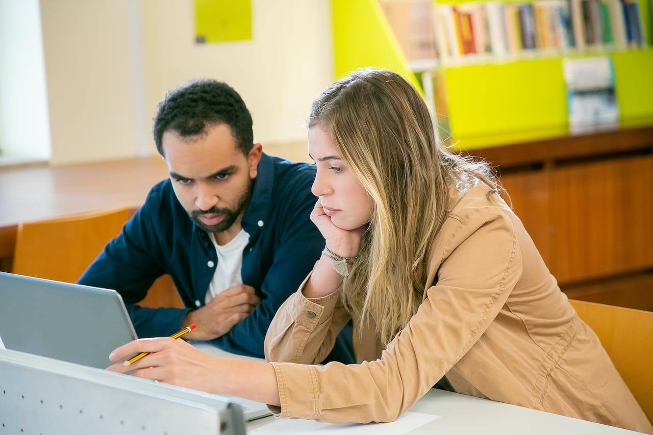 Two students collaborate at a table, one writing with a pencil while looking at a laptop, surrounded by books and a bright environment.
