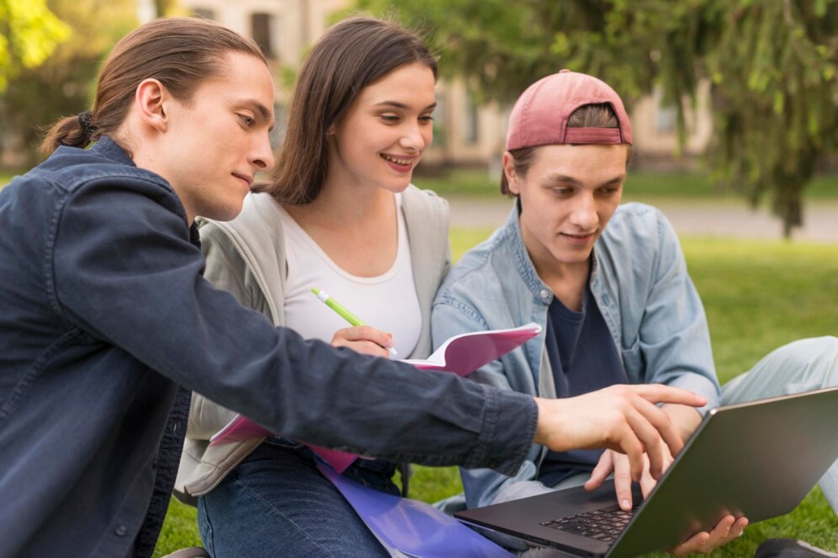 Three young adults collaborate outdoors, focusing on a laptop while taking notes in a notebook, surrounded by greenery.