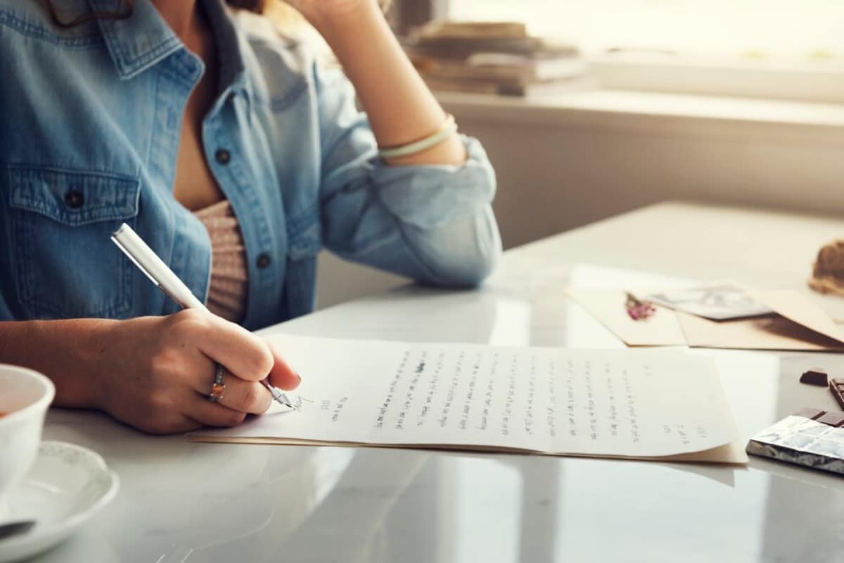 A person writes on a letter with a pen, resting their chin on their hand, while a cup of tea and photos sit on the table nearby.